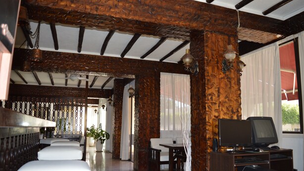 Um hall de hotel com vigas de madeira, assentos acolchoados brancos, uma televisão e uma grande janela com cortinas.