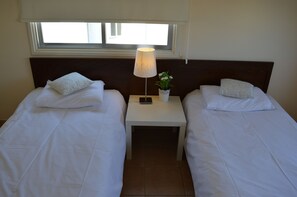 Ein Hotelzimmer mit zwei Einzelbetten, einem Nachttisch mit Lampe und einer Blumenvase sowie einem Fenster über den Betten.