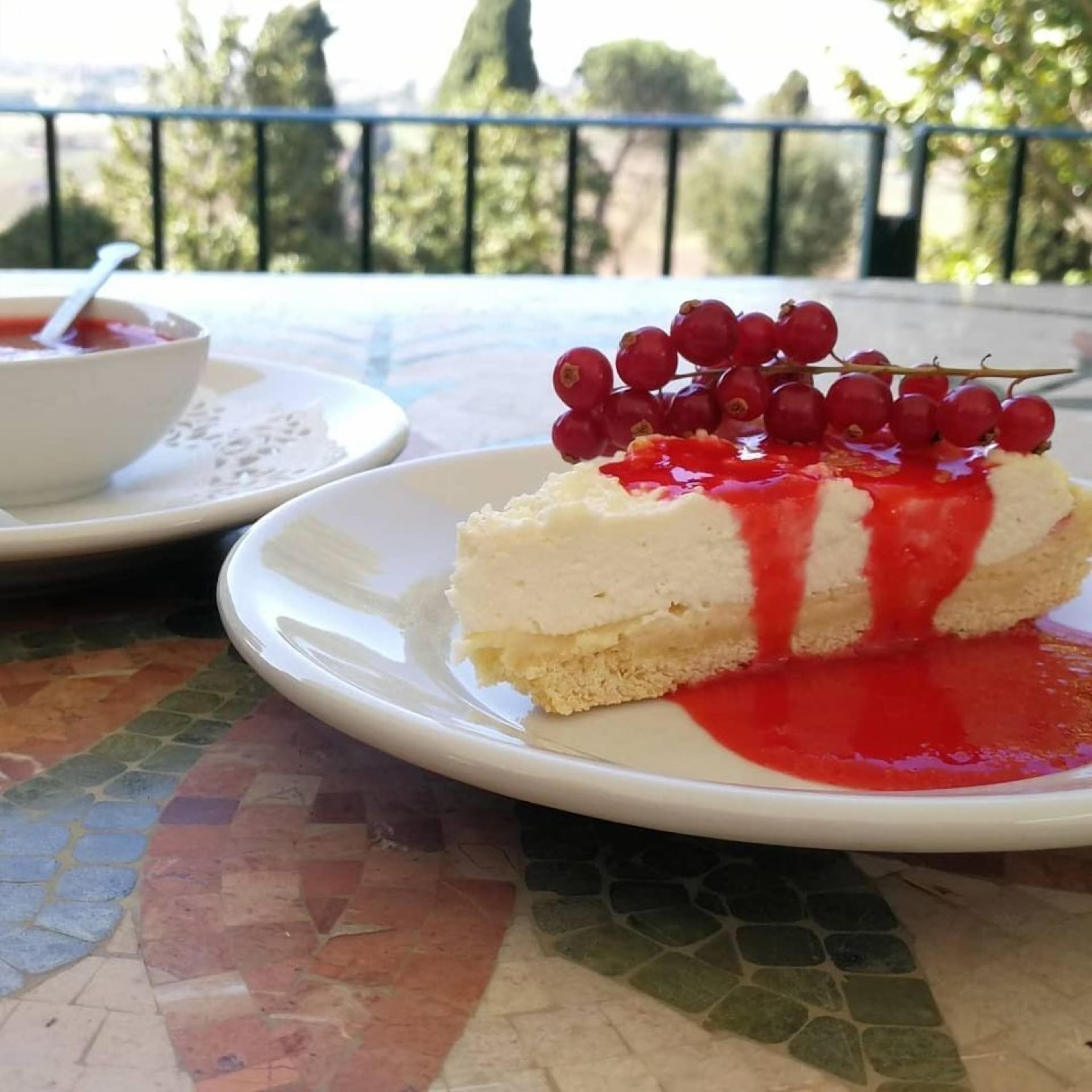 A slice of cheesecake on a white plate with red berry sauce and berries on top, placed on a mosaic table outdoors.