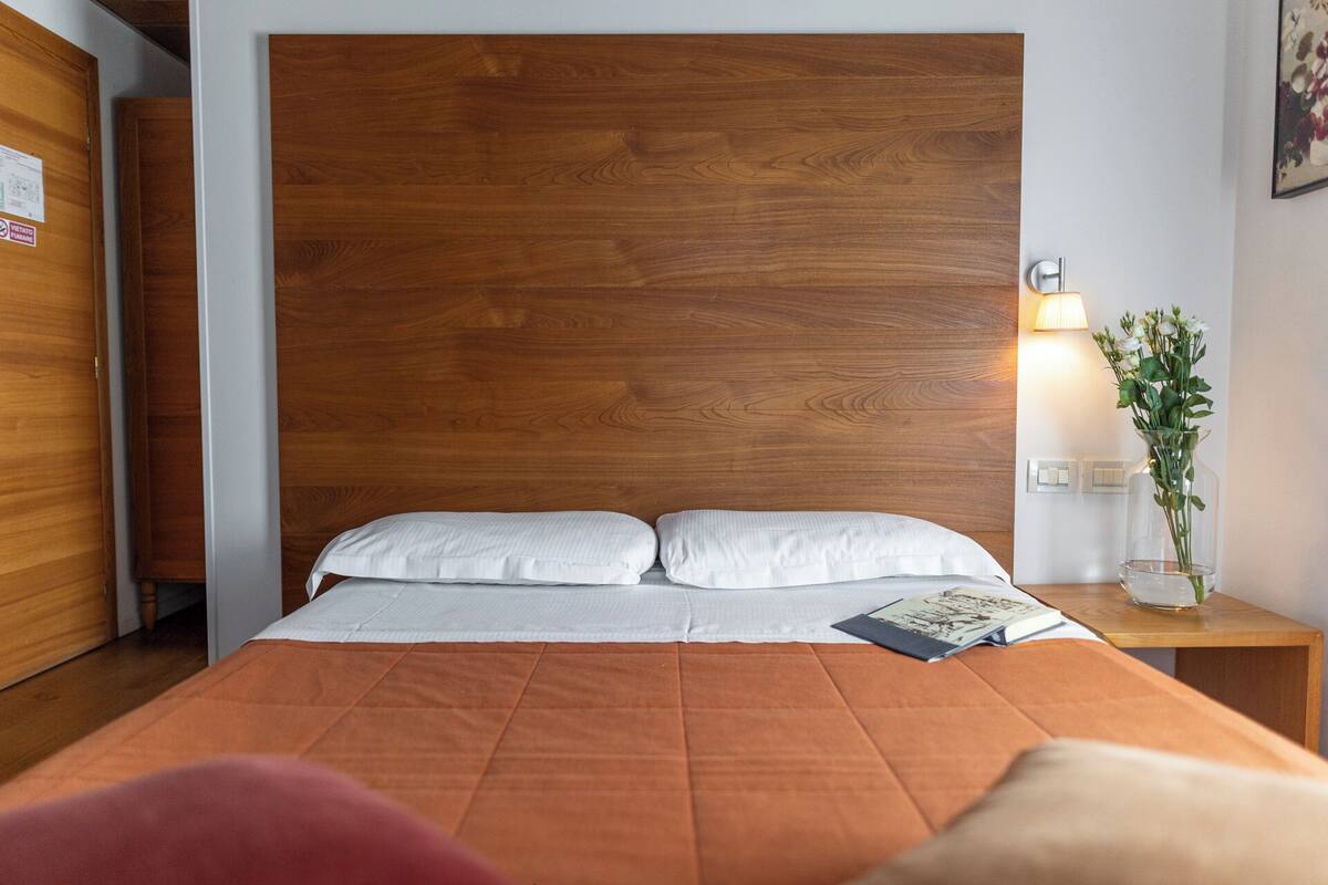 Une chambre d’hôtel avec une tête de lit en bois, un lit avec une literie blanche, un plaid orange, une table de chevet avec une lampe et un vase de fleurs, et une petite table de nuit avec un livre.
