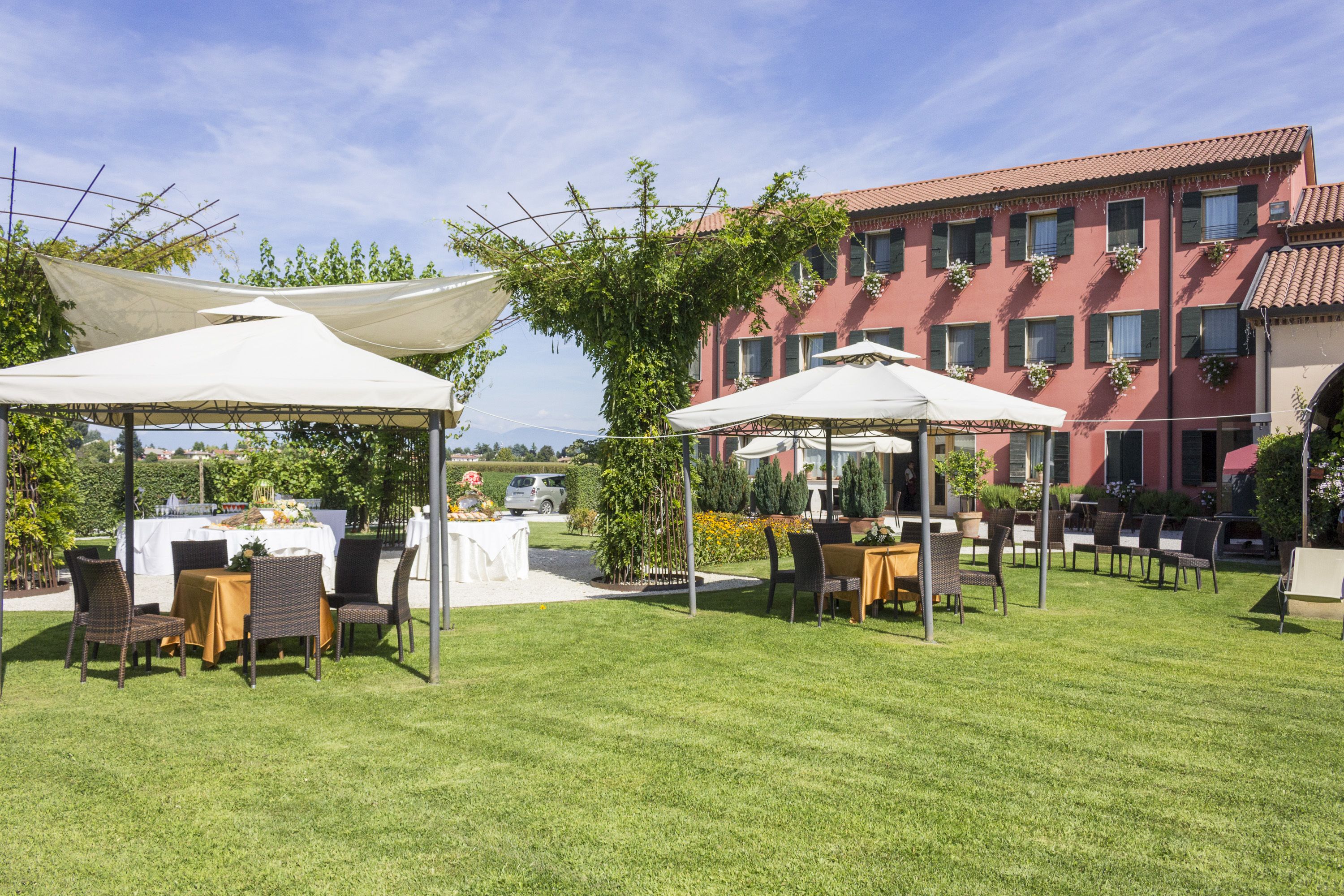 An outdoor dining area with tables and chairs under white canopies, surrounded by a well-maintained lawn and a building with multiple windows and balconies.