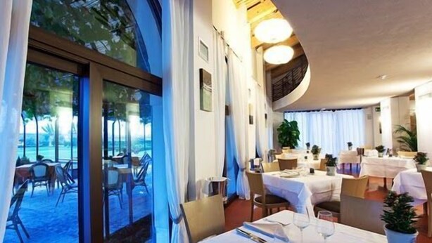 An indoor dining area with tables set for guests, large windows, and indoor plants.