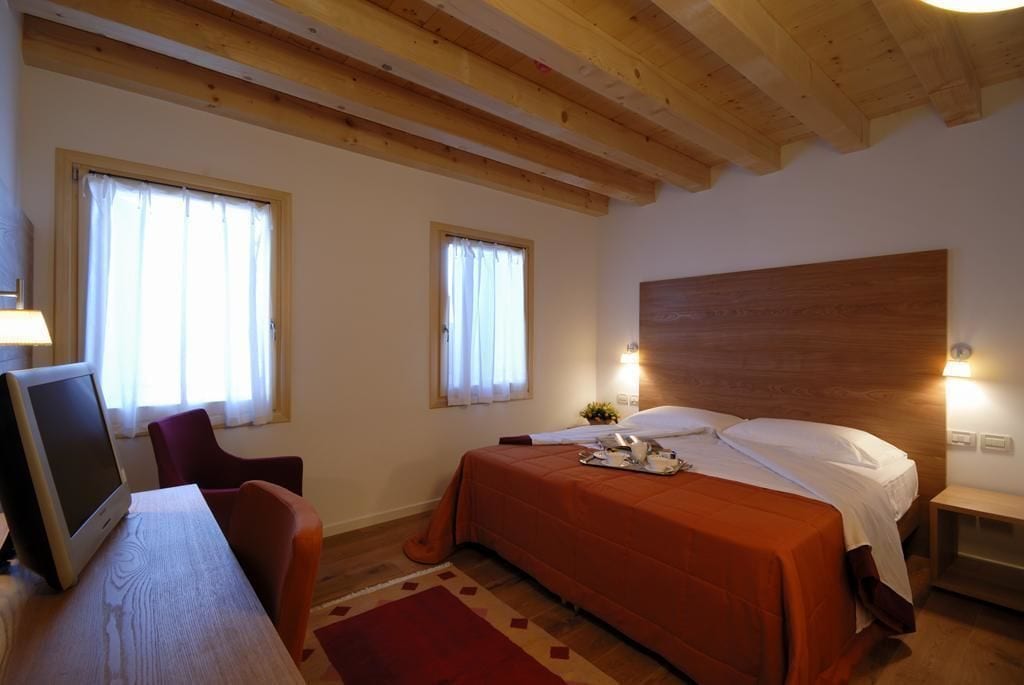 Quarto de hotel com teto de madeira, duas janelas com cortinas, uma cama com colcha laranja, mesa de cabeceira com abajur e escrivaninha com monitor de computador.