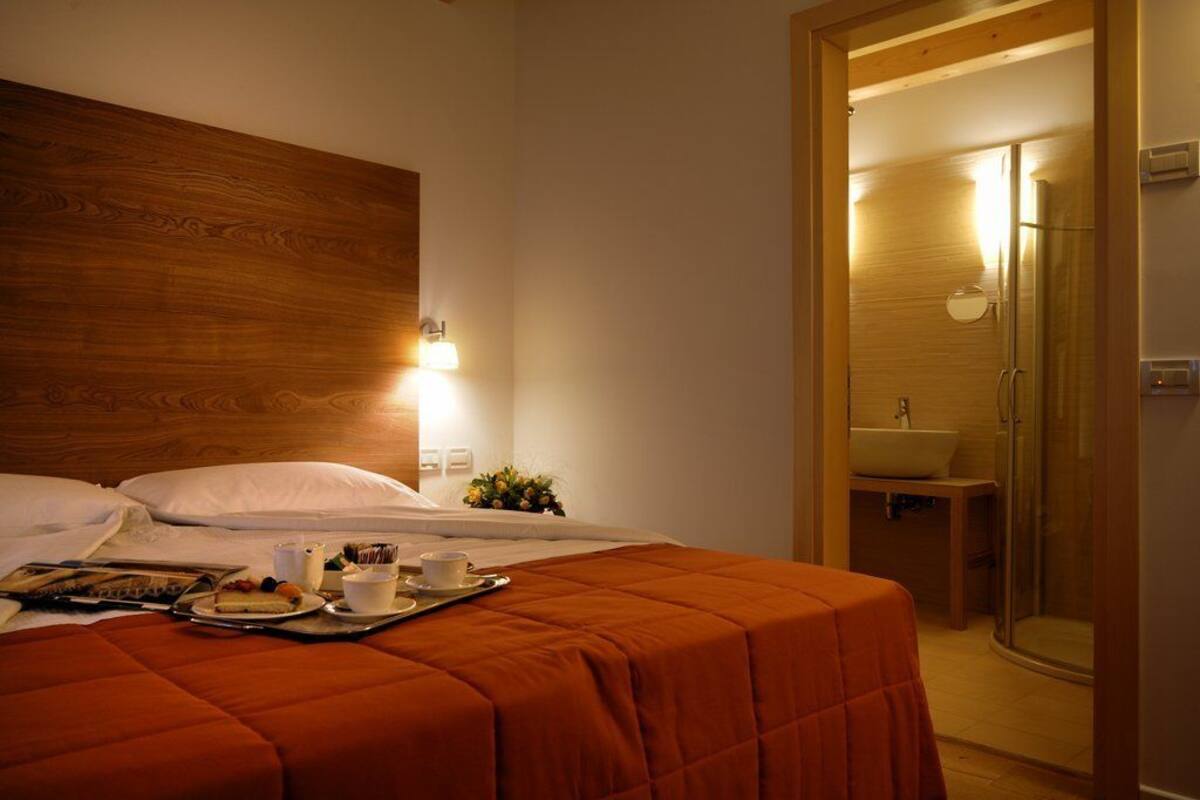 Une chambre d’hôtel avec une tête de lit en bois, un lit recouvert d’une courtepointe orange, un plateau avec des articles pour le petit-déjeuner et une salle de bain dotée d’une douche en verre.
