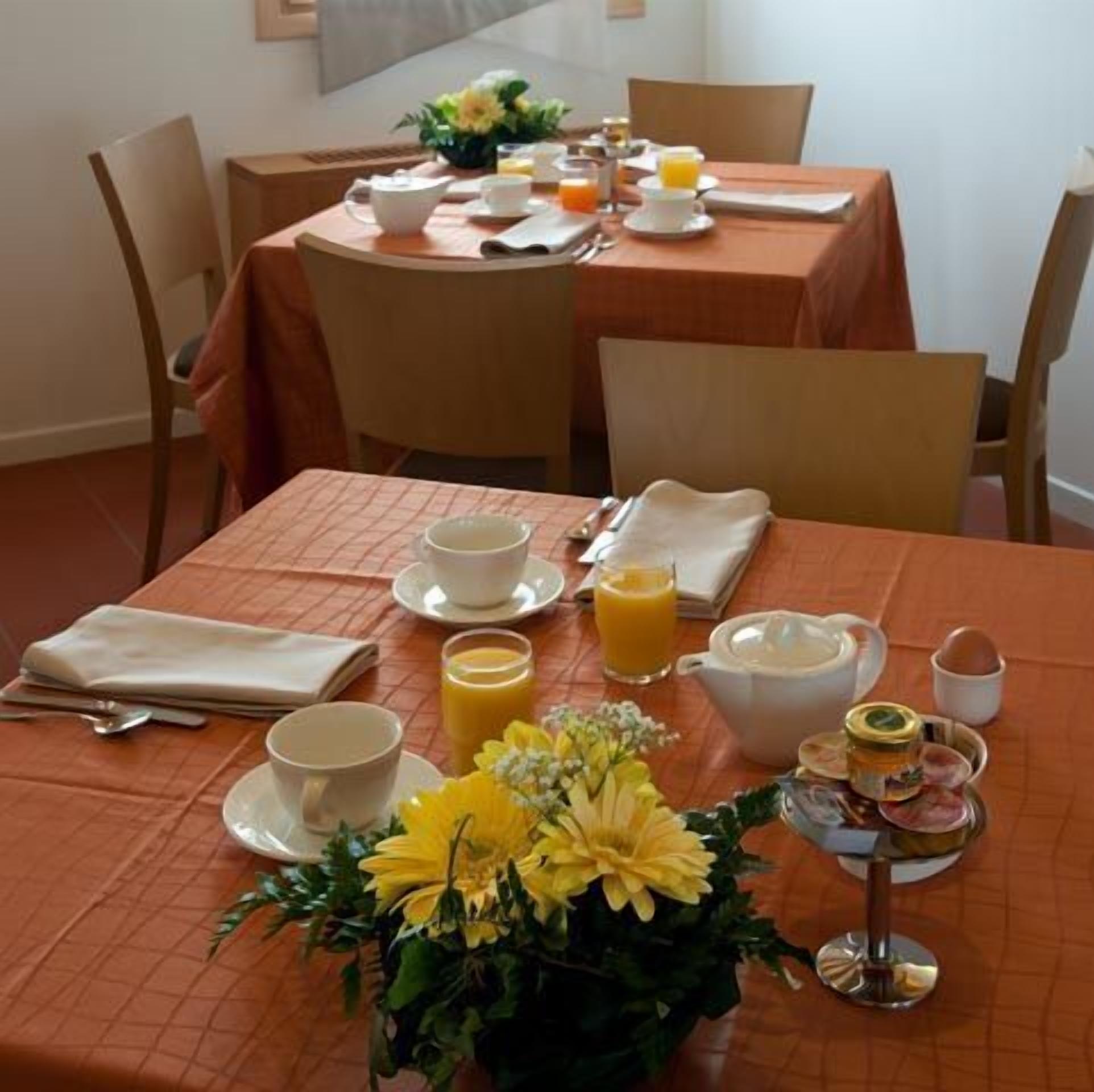 Une table mise pour le petit-déjeuner, avec une nappe pour deux personnes, comprenant des tasses, un vase de fleurs et une présentation de fruits.