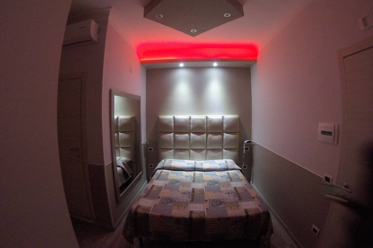 A narrow hallway leading to a bedroom with a bed, a mirror, and a red light fixture above the bed.