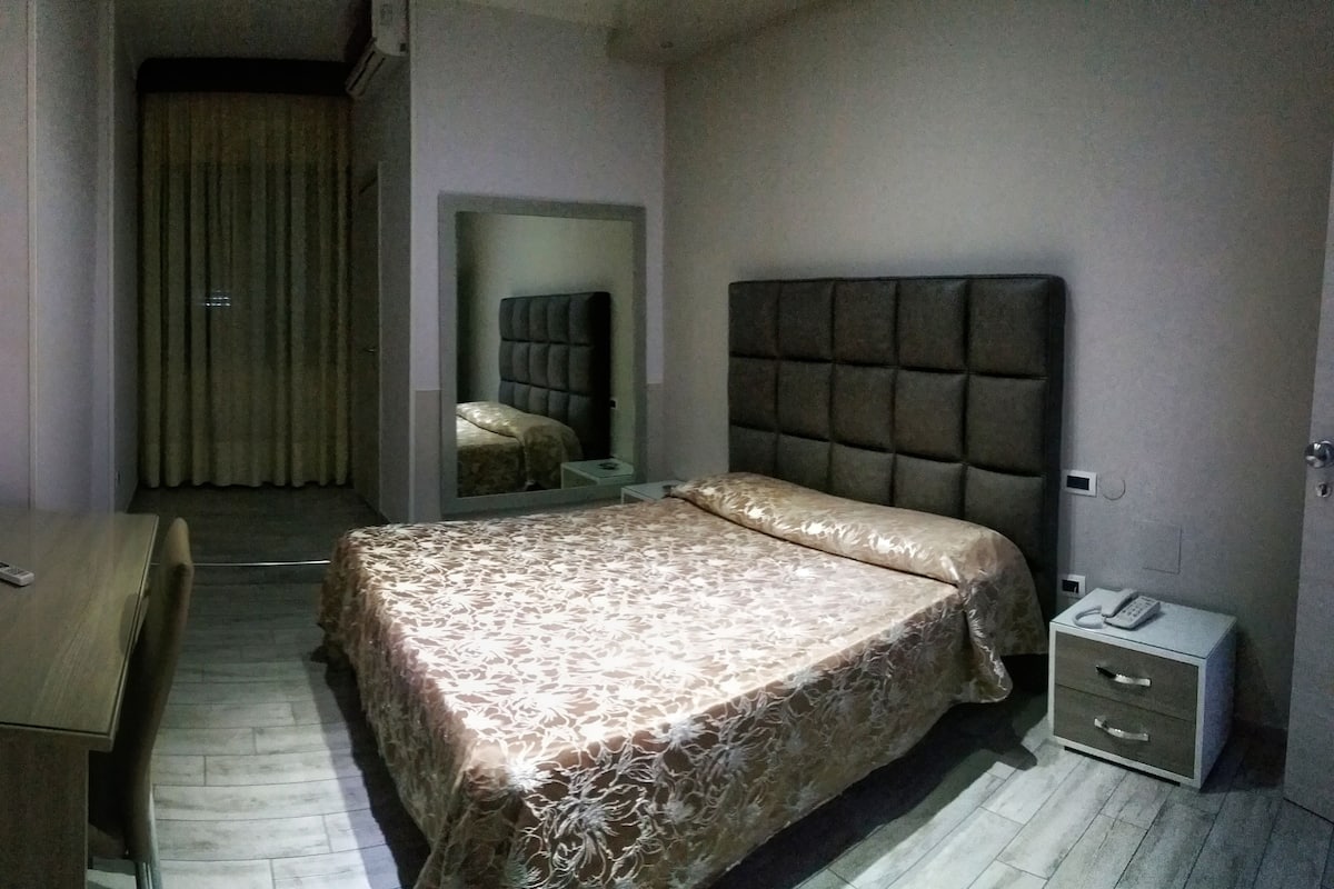 A bedroom with a bed, a bedside table, a nightstand, a mirror, and a door.