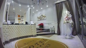 A hotel reception area with a tufted white counter, a chandelier, and a reception desk. There is a staircase leading to another level, and a large floral arrangement on the right. The floor has a circular logo with the hotel's name.