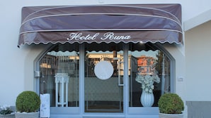 The exterior of Hotel Runa with a large awning, a glass entrance, and two potted plants on either side.
