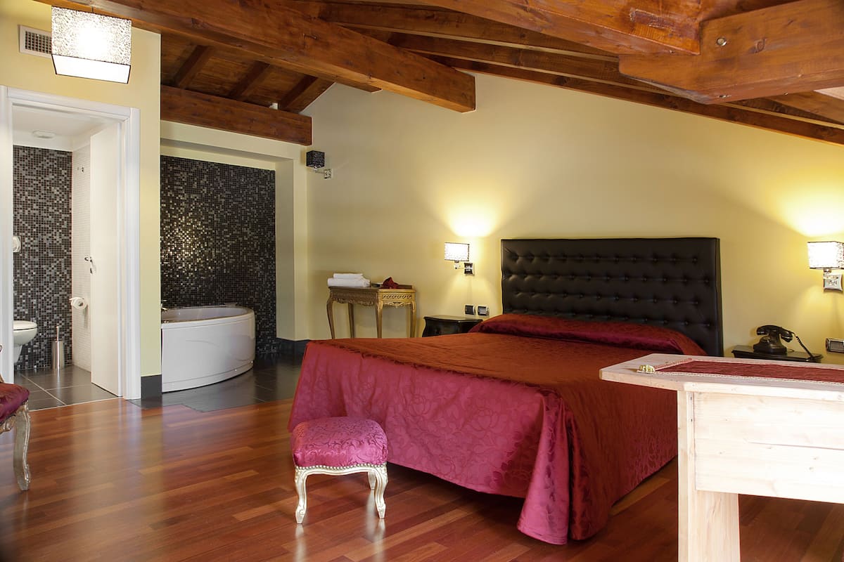 A bedroom with a large bed, a wooden ceiling, a nightstand, a lamp, and a bathroom visible through an open door.