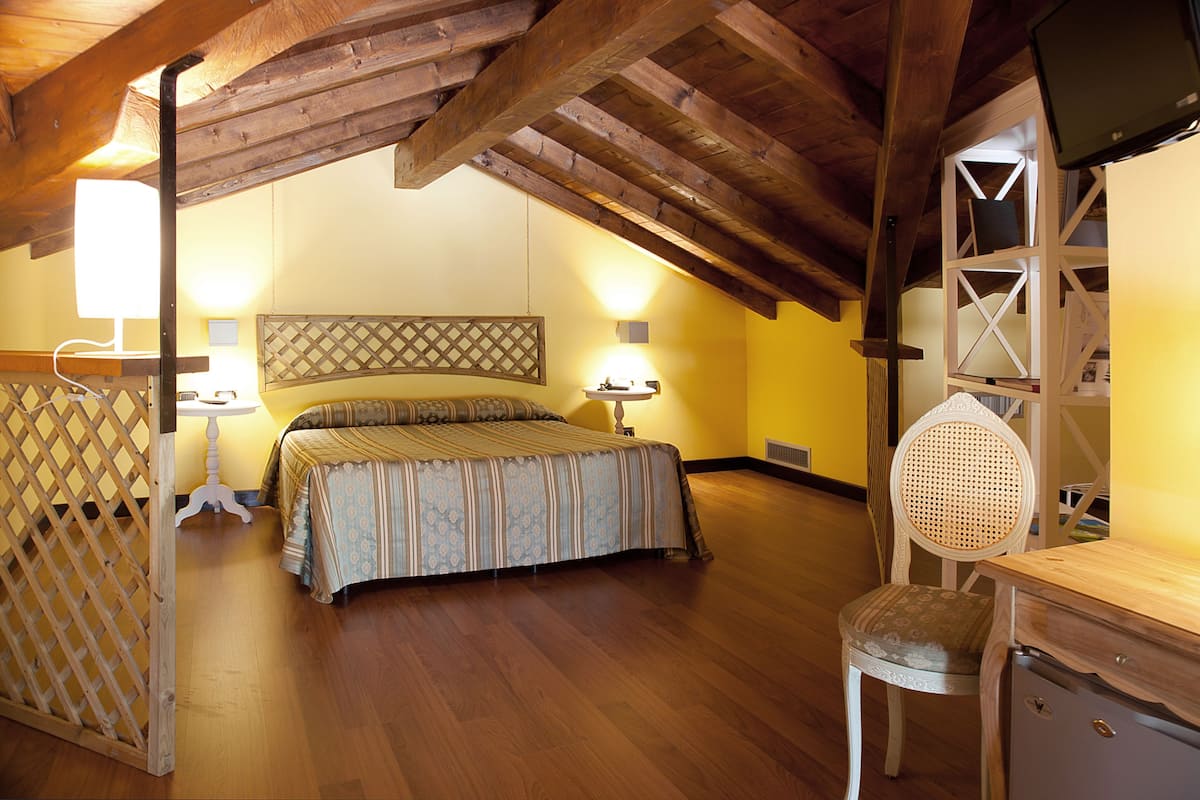 A bedroom with a wooden ceiling, a bed with a patterned bedspread, a nightstand with a lamp, and a chair with a woven seat.