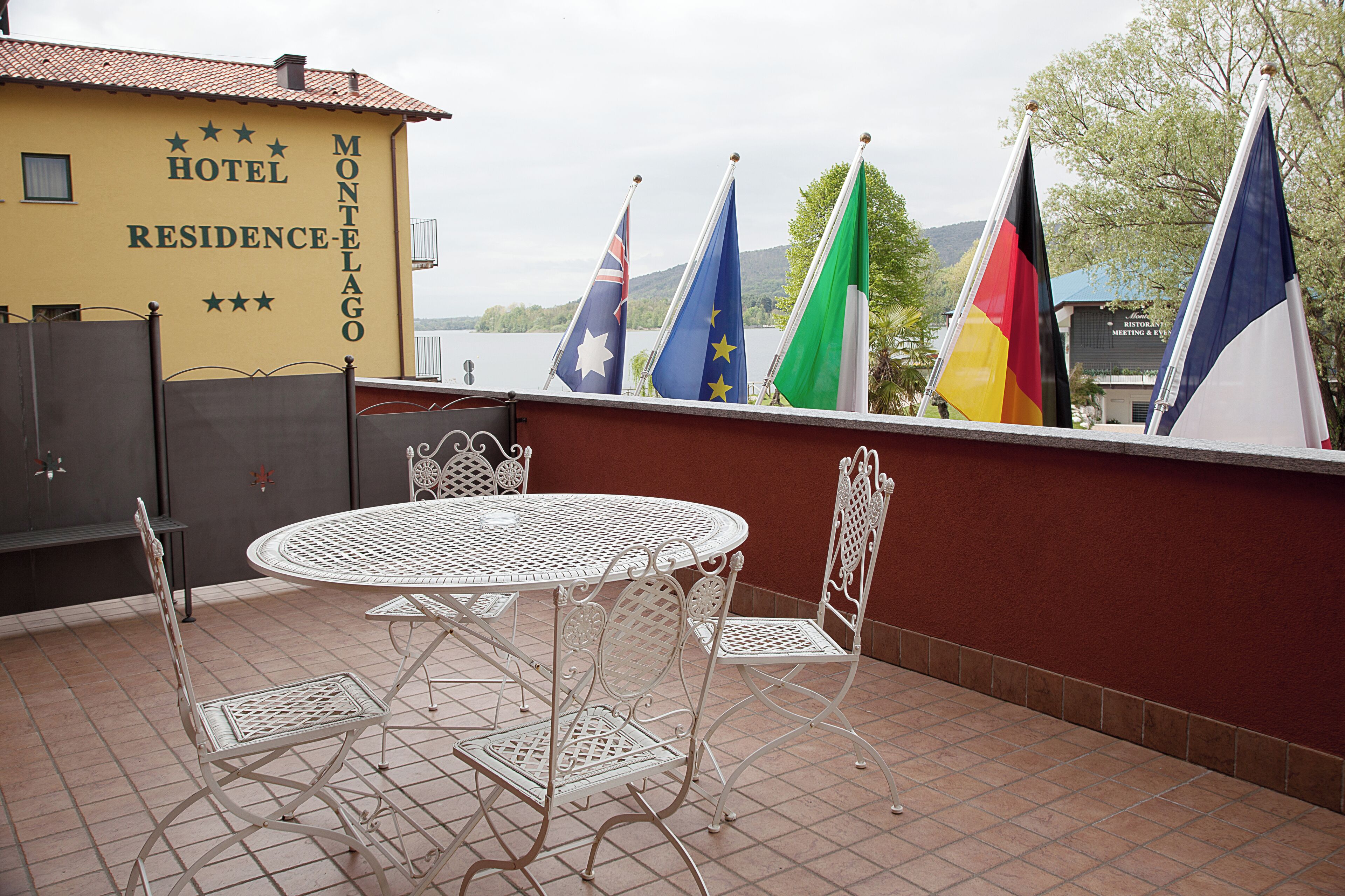 Une terrasse avec une table ronde en métal et des chaises, un bâtiment jaune portant l’inscription « HÔTEL-RÉSIDENCE », et des drapeaux de différents pays.