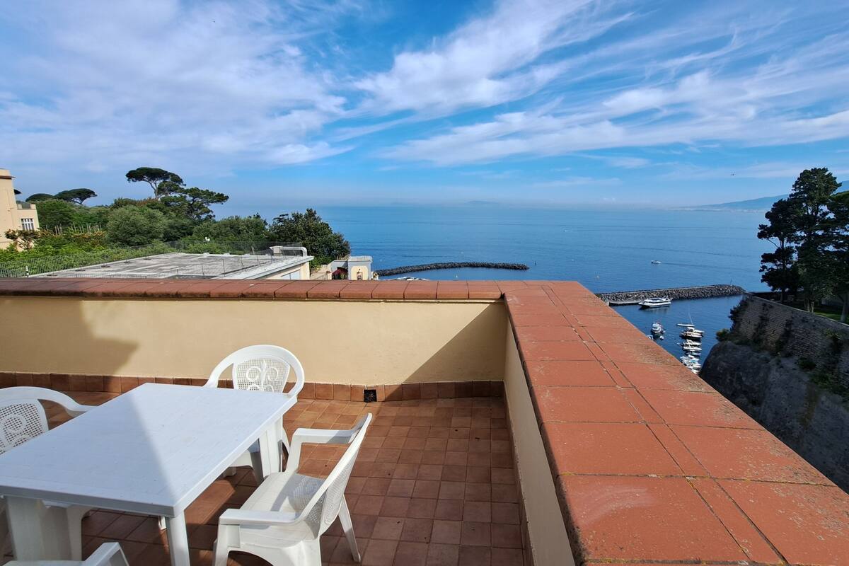 A terrace with a table and chairs, overlooking a coastal area with boats and a clear sky.