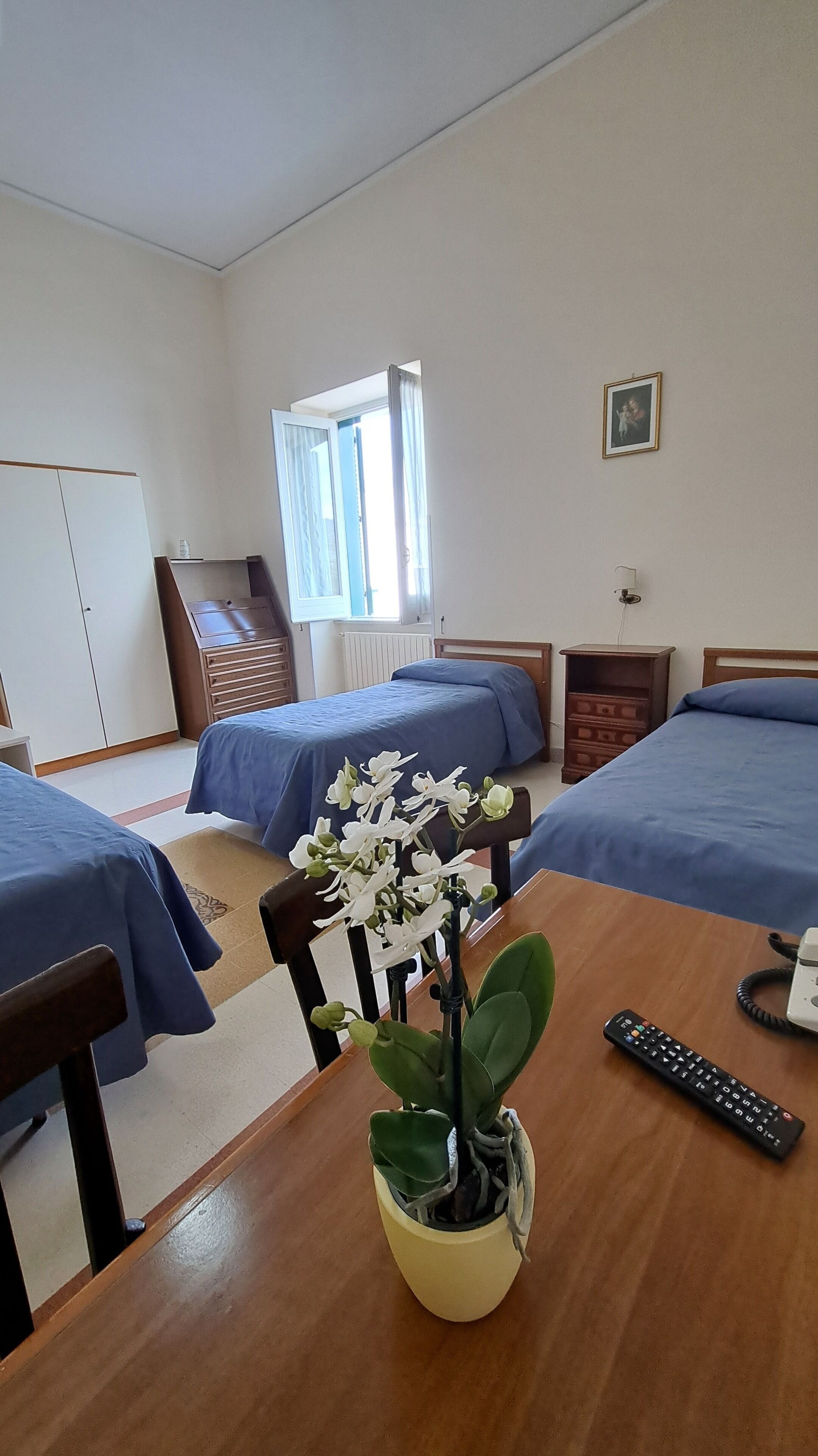Una camera d'albergo con due letti, un tavolo di legno, un'orchidea in vaso, un telecomando e una finestra con tende.