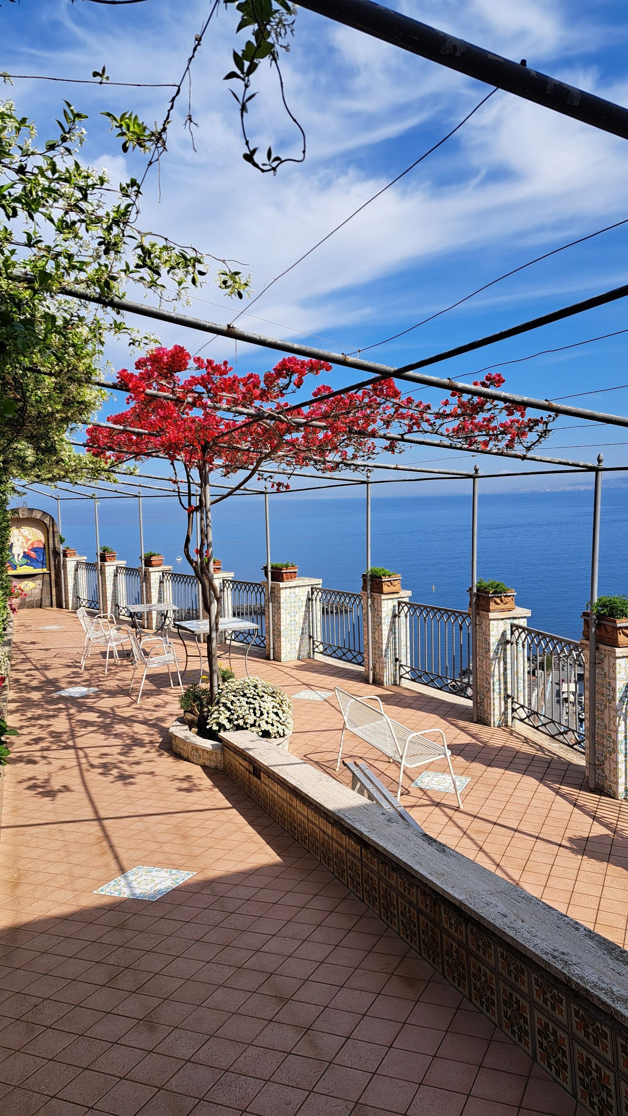 A terrace with tiled flooring, metal chairs, and a railing overlooking a body of water.