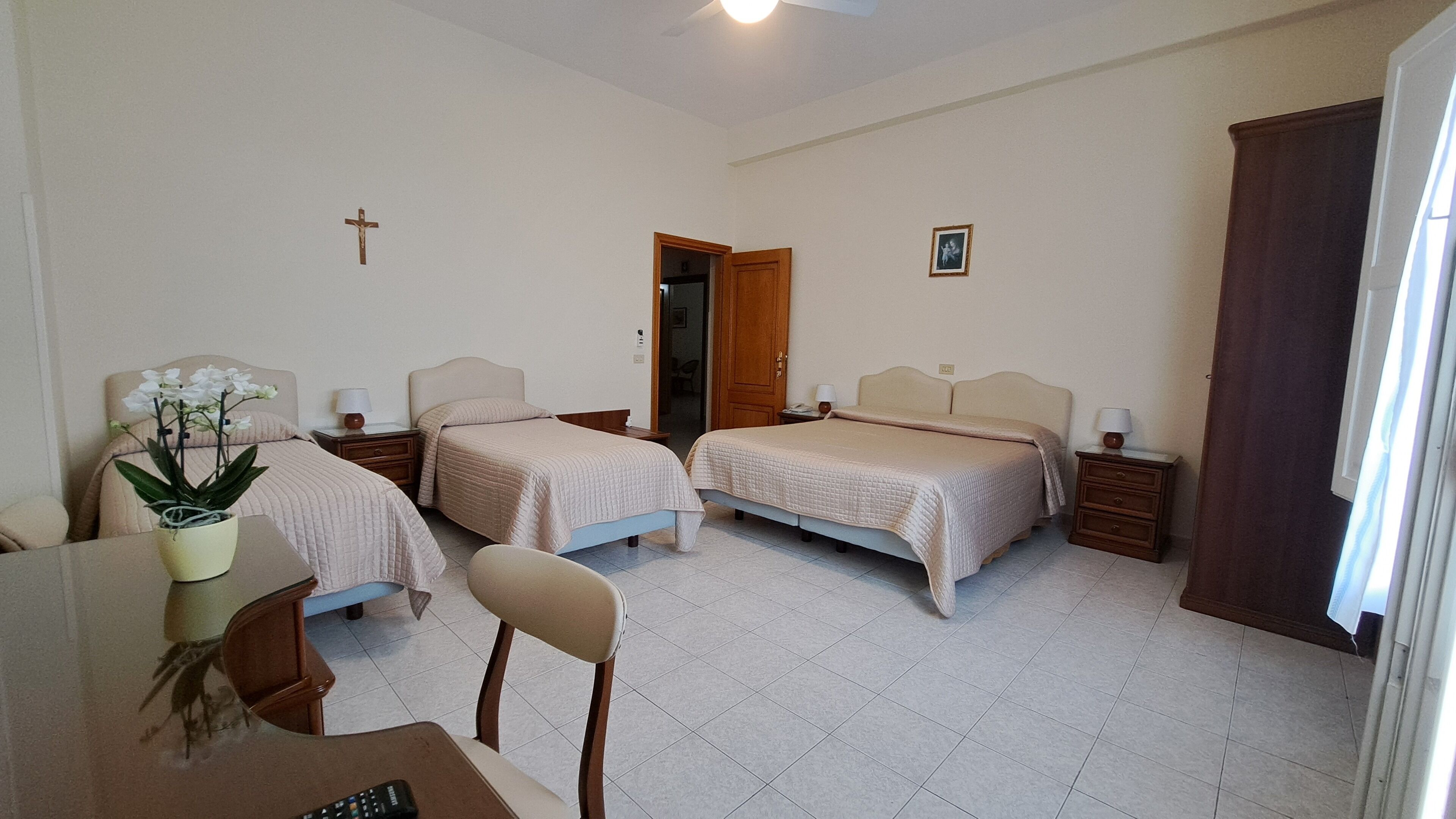 A hotel room with two beds, a small table with a plant, a chair, a cross on the wall, and a door leading to another room.