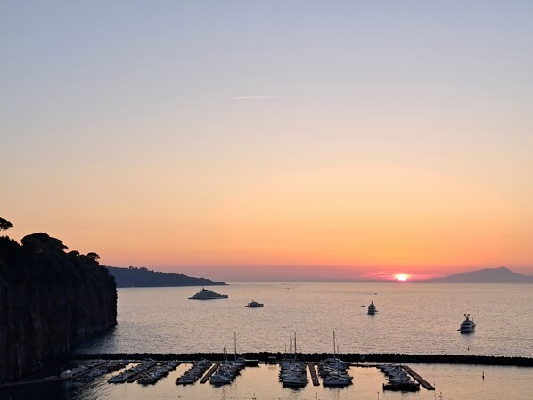 A marina with boats docked at the pier, a sunset in the background, and a rocky coastline.