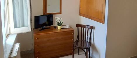 Una habitación pequeña y poco amueblada con un aparador de madera, una silla, un escritorio con una computadora, un espejo y un radiador.