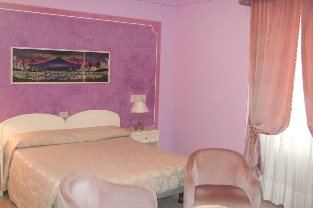 A bedroom with a bed, two chairs, a nightstand, a lamp, and a painting on the wall.