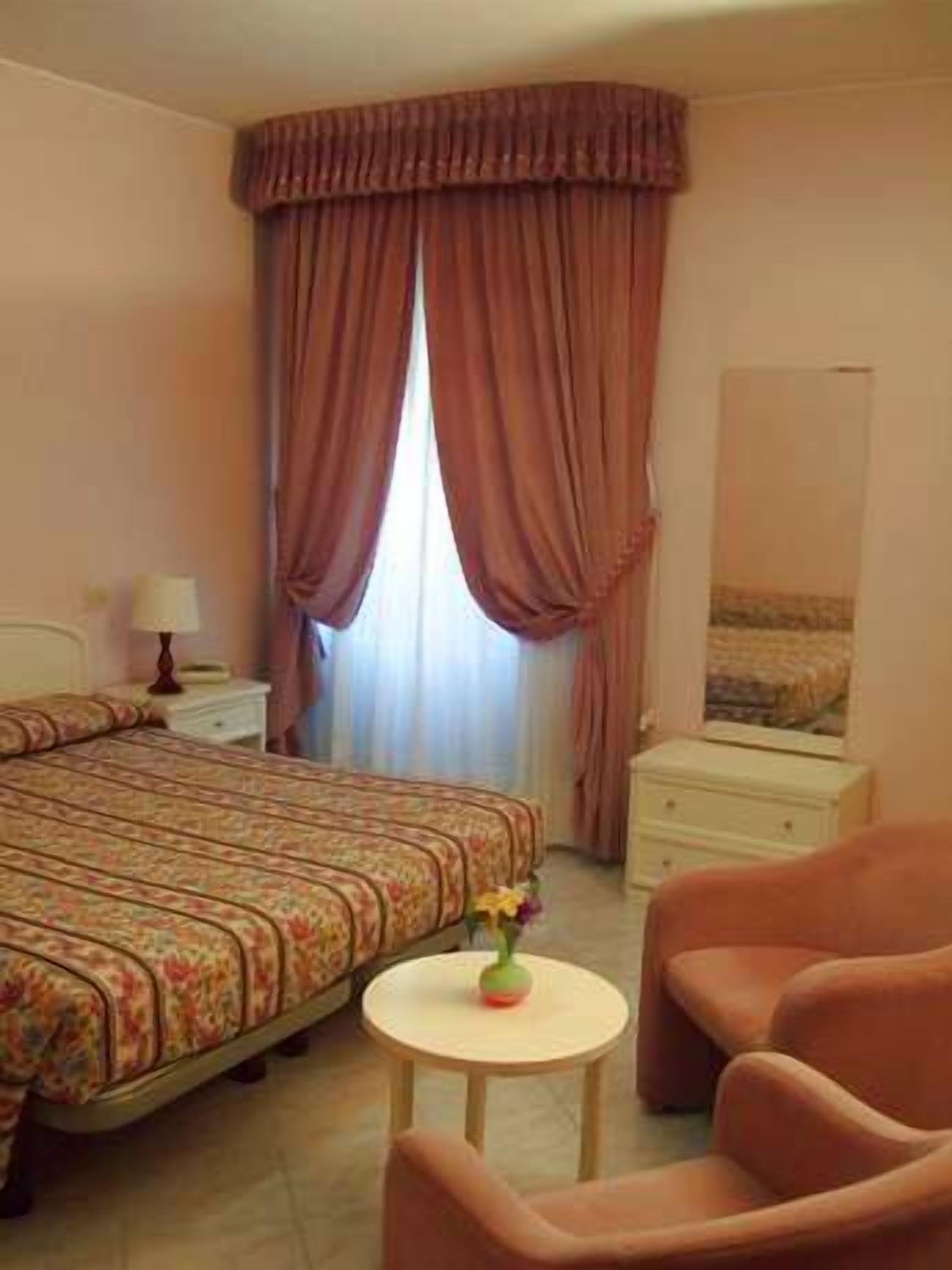 Une chambre d’hôtel comprenant un lit, une table de chevet avec une lampe, une petite table ronde avec un vase et une chaise.