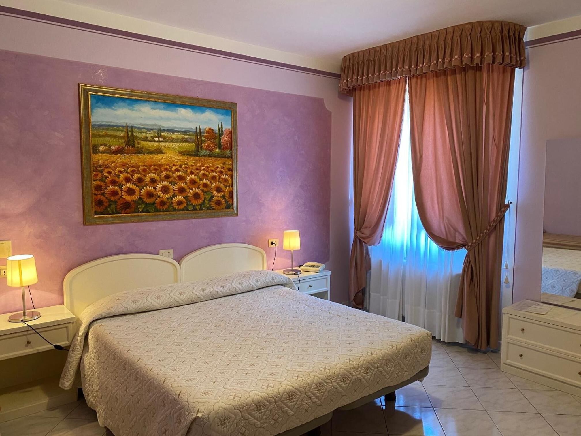 A bedroom with a bed, nightstands, a lamp, a painting, and a window with curtains.