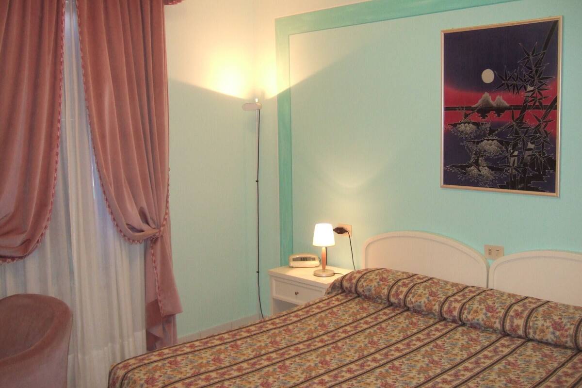 A bedroom with a bed, a nightstand, a lamp, a painting, and a window with curtains.