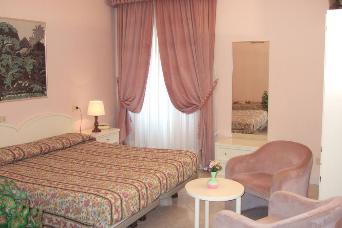 A bedroom with a bed, a bedside table with a lamp, a chair, a small table with a vase, and a mirror on the wall.