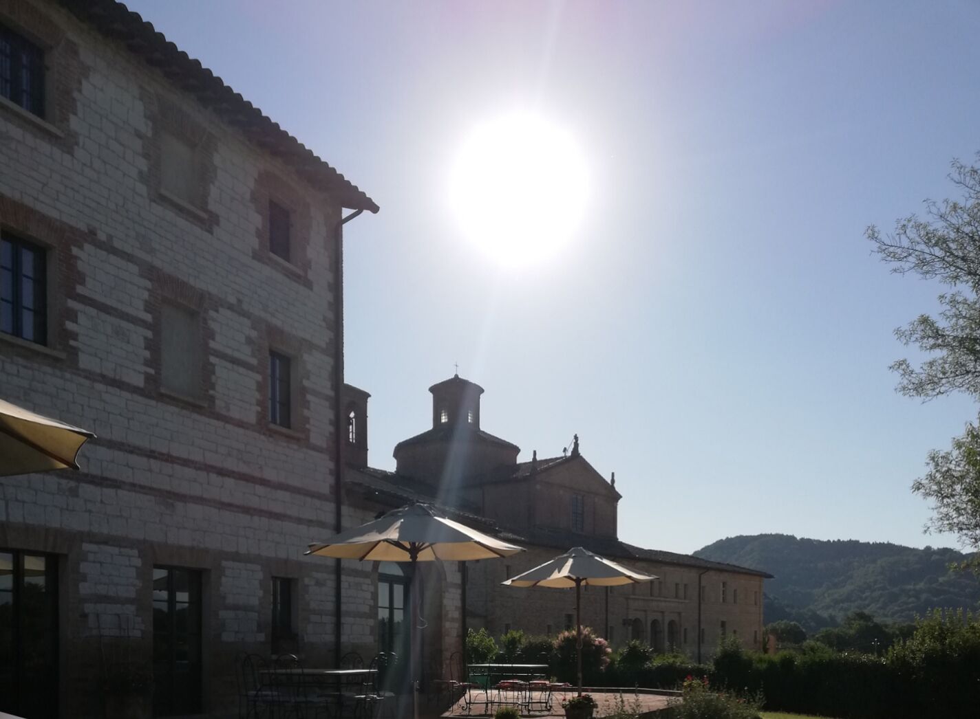 Un edificio con fachada de ladrillo y un campanario, asientos exteriores con sombrillas y un cielo despejado.
