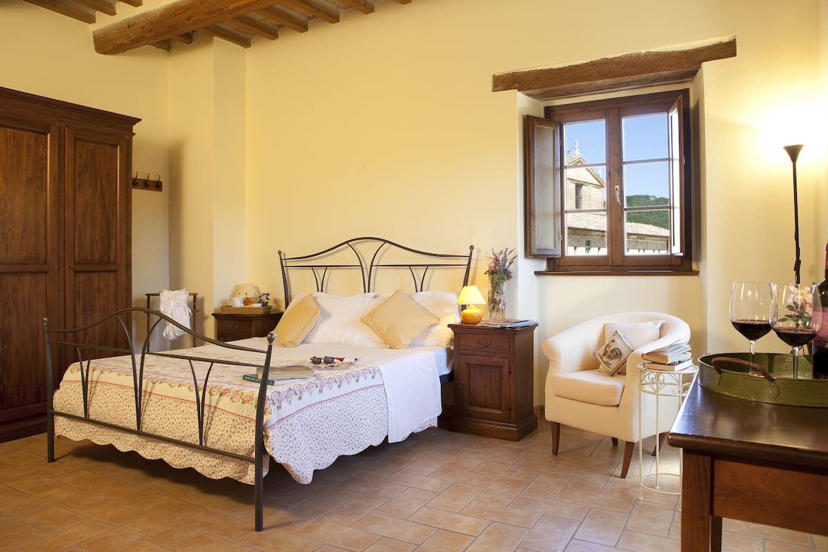 A bedroom with a bed, a nightstand, a chair, a window, and a table with glasses.