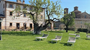 A courtyard with a stone building, a large tree, and several lounge chairs. - Country House Parco Ducale (Urbania)