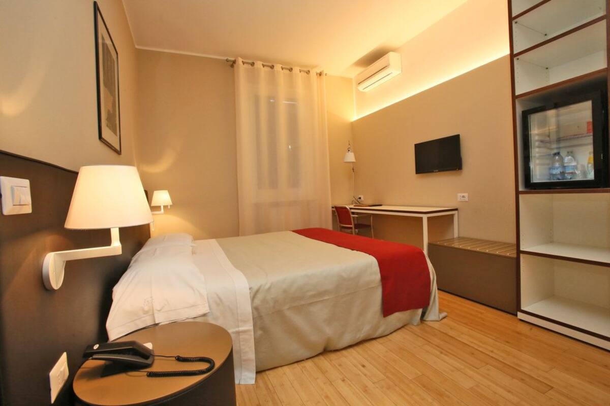 A hotel room with a bed, bedside table, lamp, desk, chair, and a minibar.