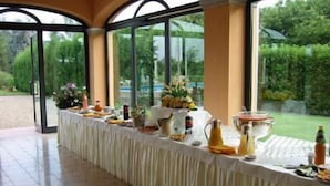 Un'area pranzo all'aperto riparata, con un lungo tavolo apparecchiato per un pasto, circondata da pannelli di vetro e colonne.