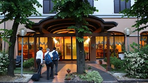 L'ingresso di un hotel con una tettoia, piante in vaso e un interno ben illuminato visibile attraverso le vetrate.