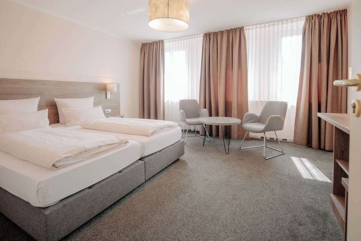 A hotel room with a double bed, a small round table, two chairs, a desk, and a lamp.