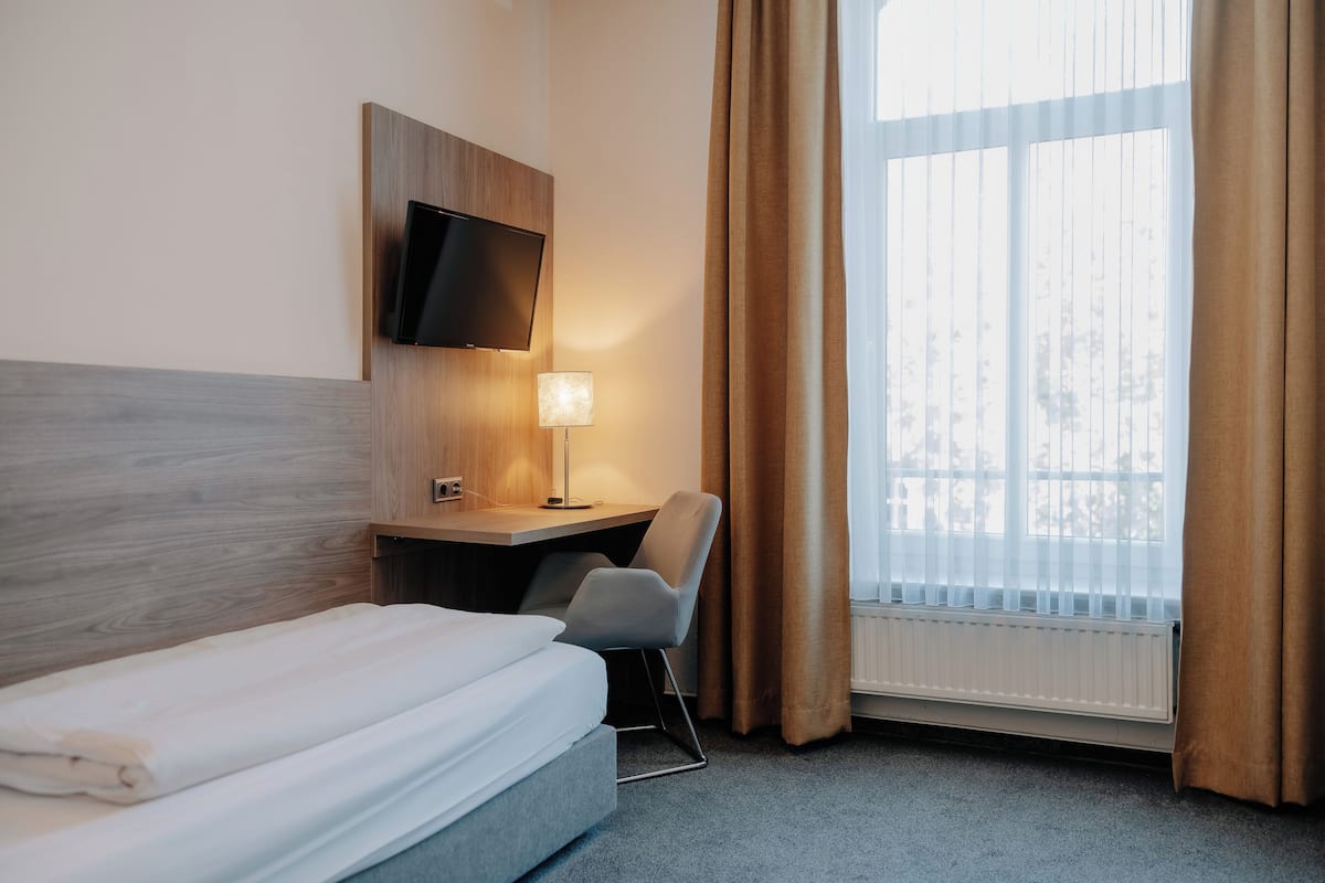 A hotel room with a bed, a desk with a chair, a TV mounted on the wall, a lamp, and a window with curtains.