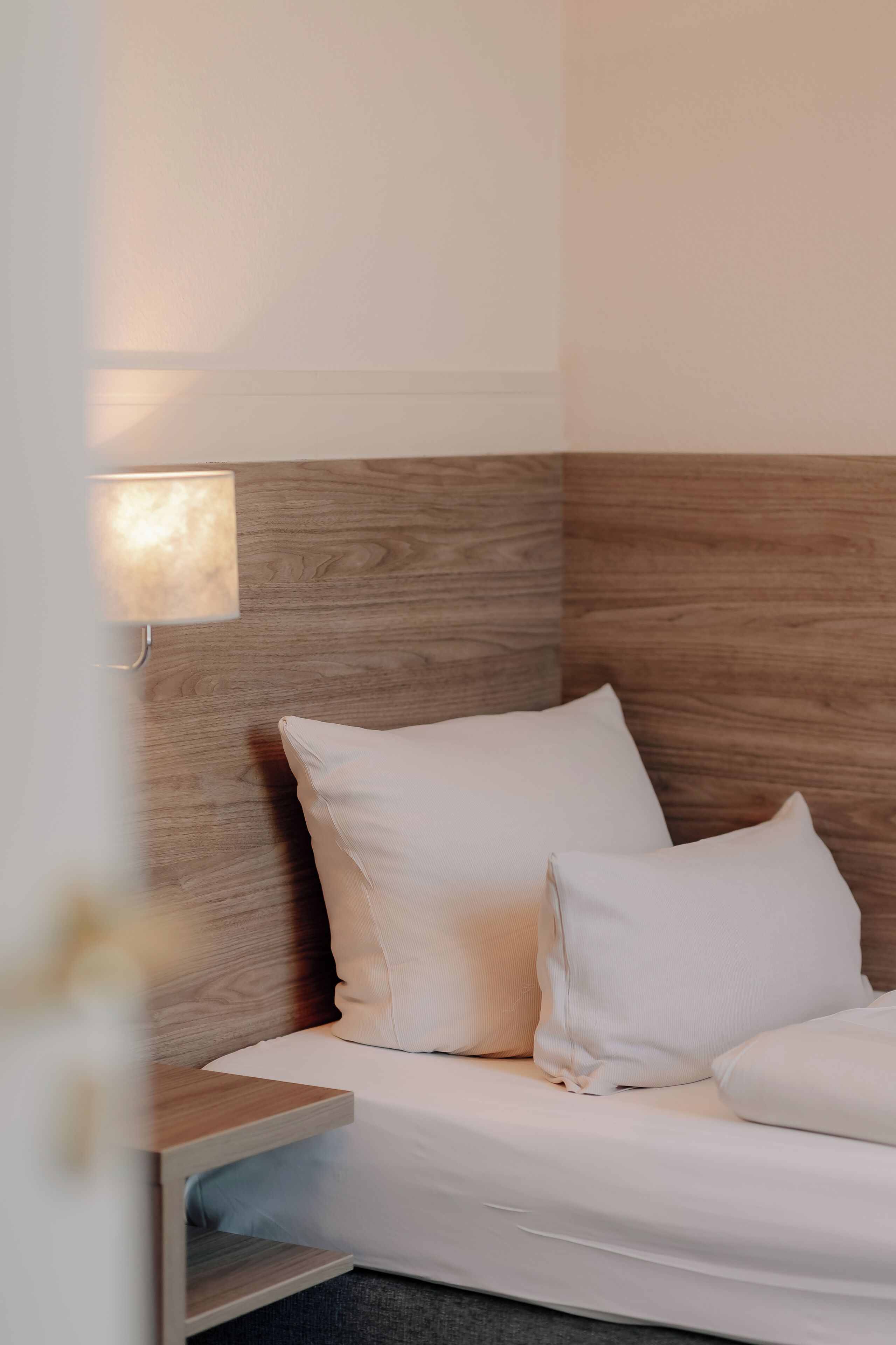 Quarto de hotel com cama, almofadas brancas, mesa de cabeceira e luminária fixada na parede.