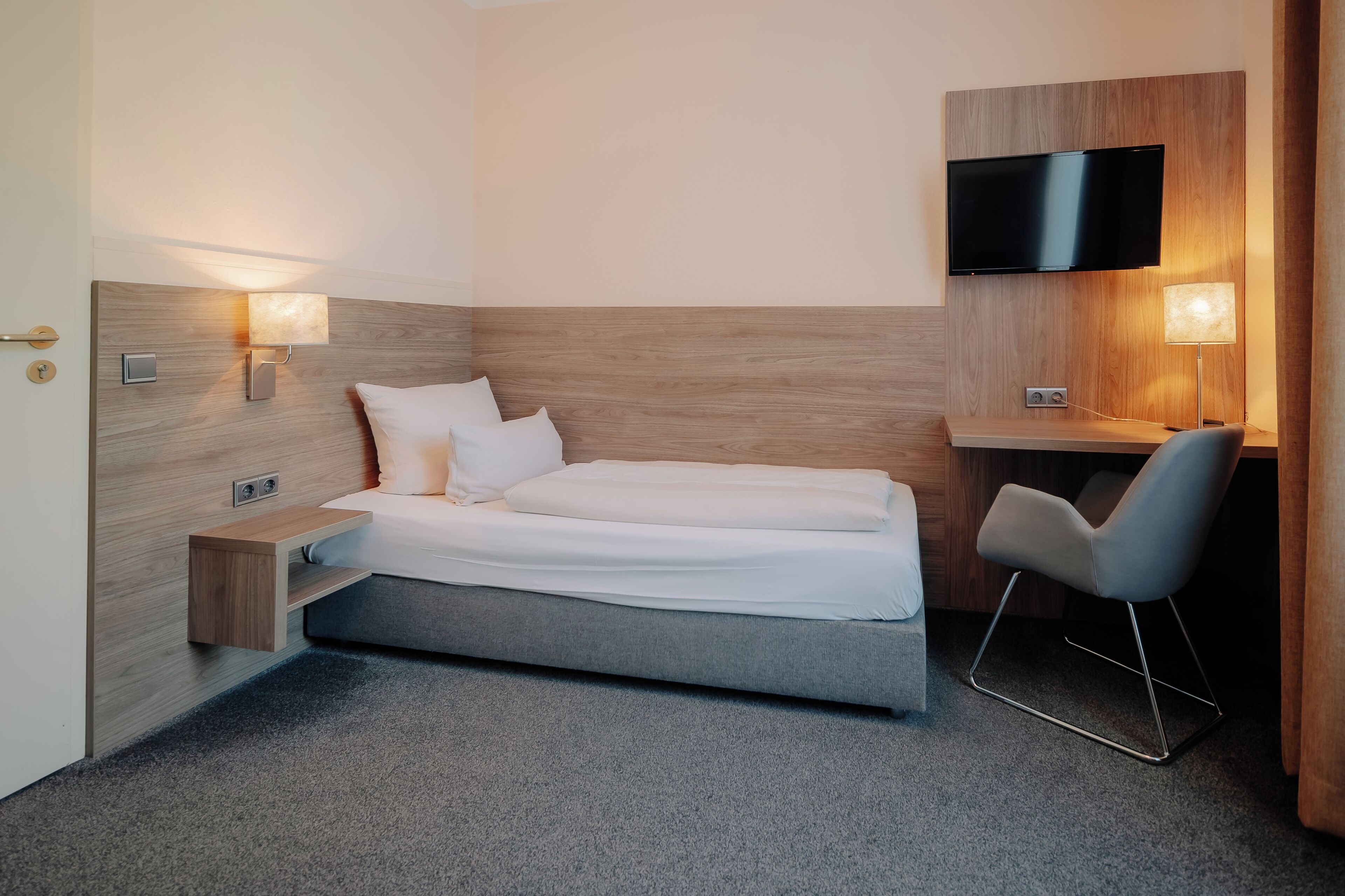 Ein modernes Hotelzimmer mit Bett, Nachttischen, einem Schreibtisch, einem Stuhl und einem an der Wand befestigten Flachbildfernseher.