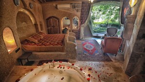 A room with a bed, a sitting area, a bathtub with rose petals, and a view of greenery outside. - Traveller's Cave Hotel (Nevsehir)