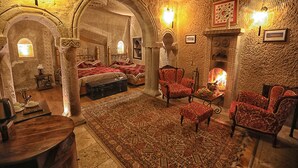 A room with a fireplace, red upholstered chairs, a patterned rug, and a bed with a red bedspread. - Traveller's Cave Hotel (Nevsehir)