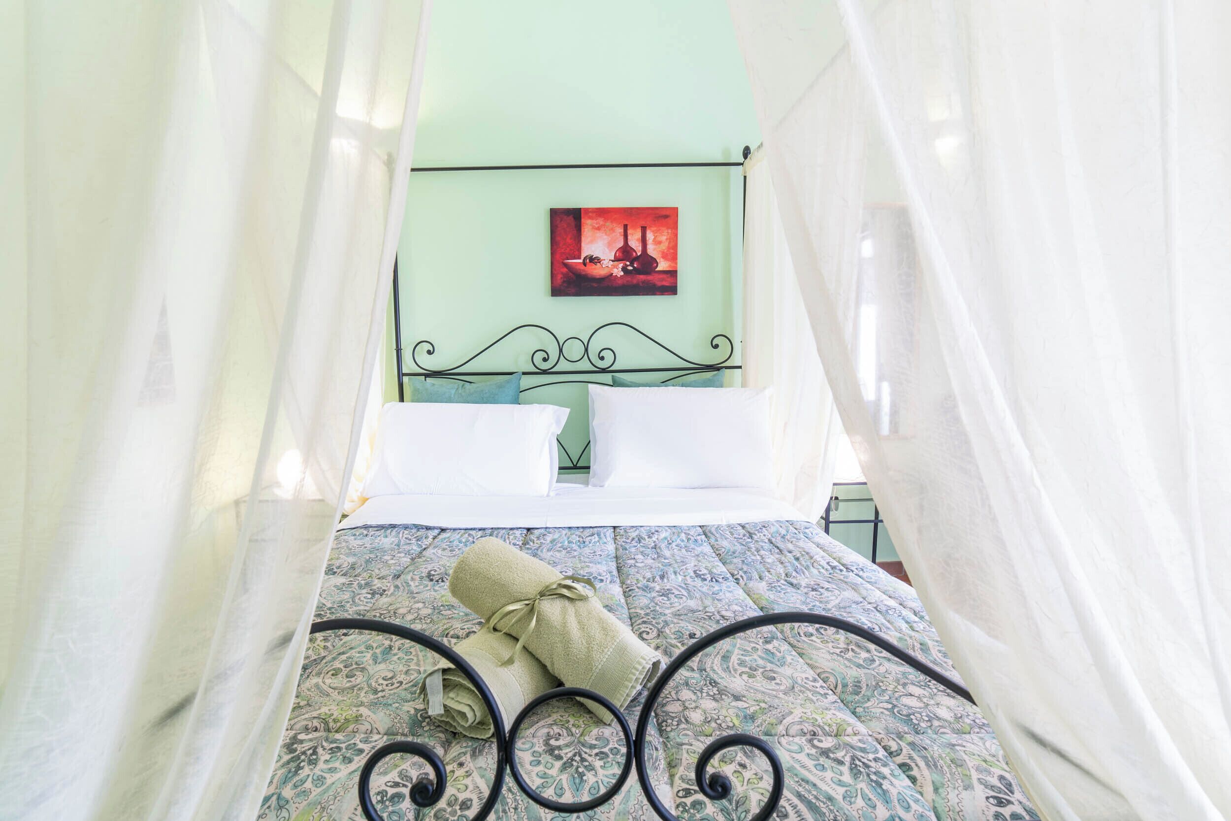 A bed with a patterned bedspread, white pillows, and a rolled towel. A painting is on the wall behind the bed.