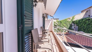 A balcony with wicker chairs and a table, a green door, and a view of a building with a red-tiled roof and greenery.