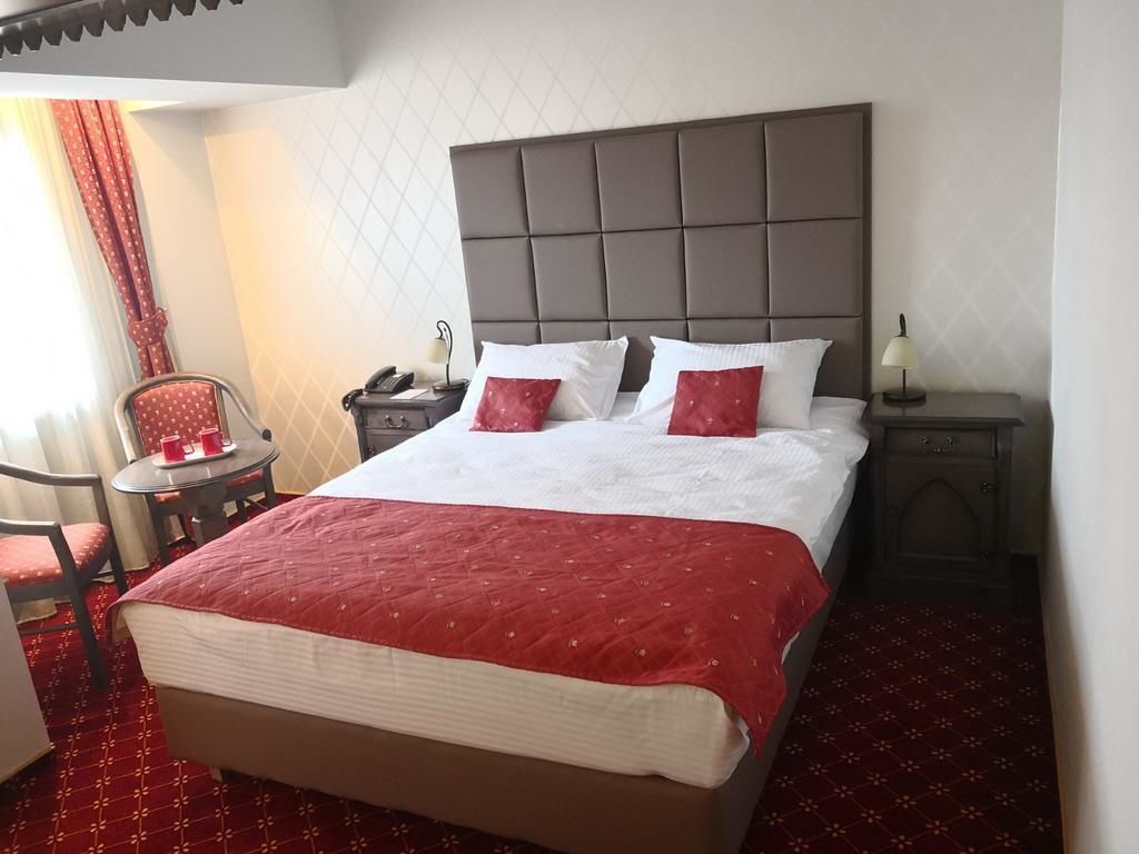 A hotel room with a large bed, a bedside table with a lamp, a chair, and a small table with a phone and a cup.