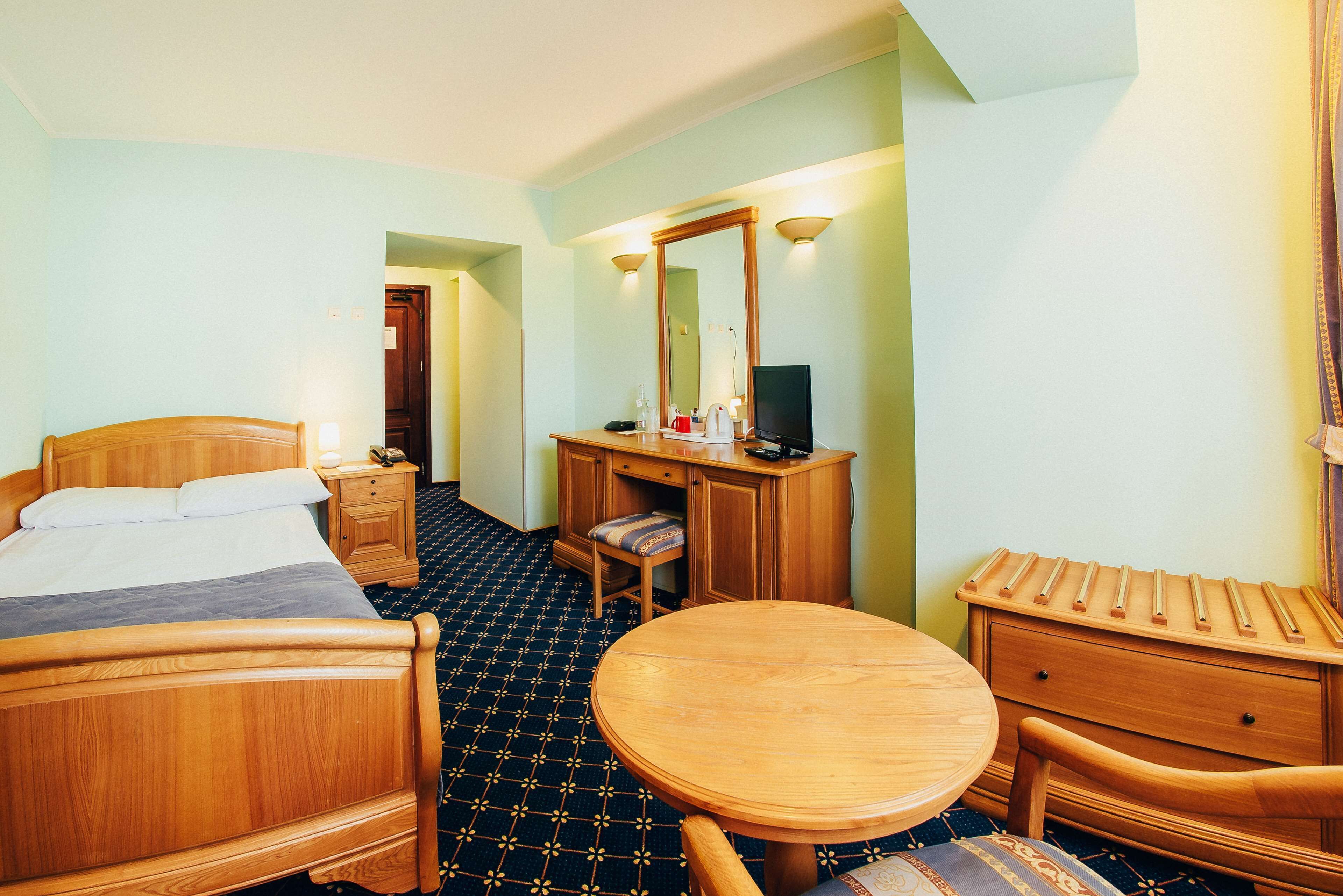 Habitación de hotel con una cama de madera, un escritorio con silla, una mesa redonda de madera y un aparador.