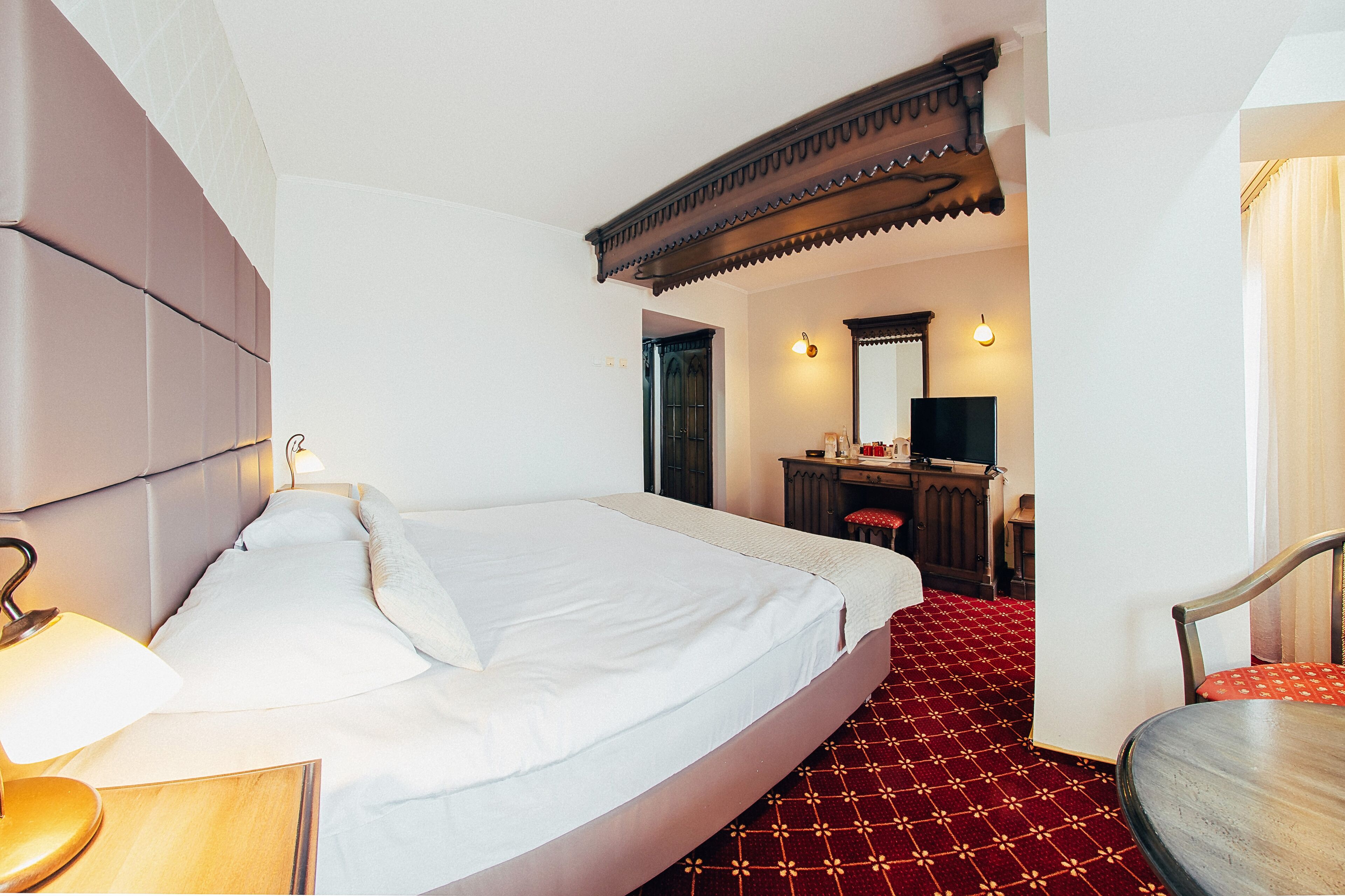 Habitación de hotel con una cama grande, un escritorio con silla, un espejo y una alfombra roja estampada.