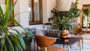 Un salon confortable avec un canapé, des fauteuils et une table basse. On y trouve des plantes en pot et un luminaire suspendu.