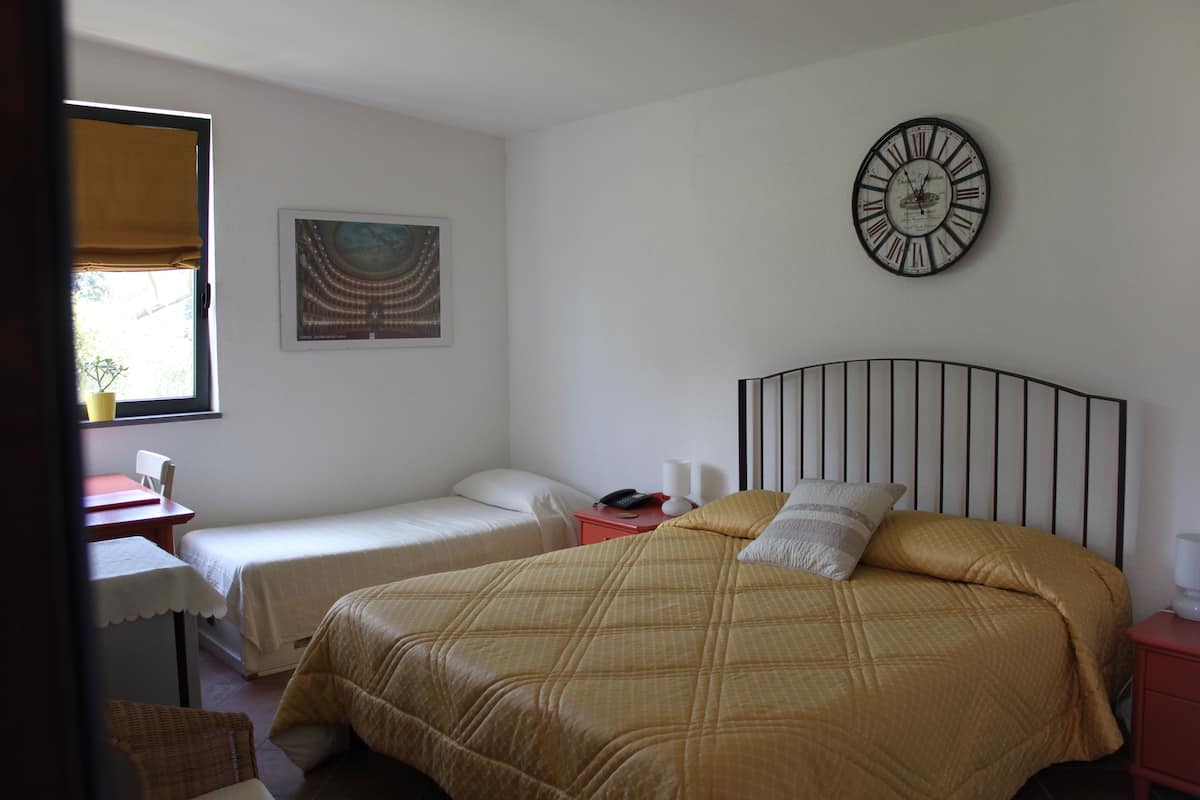 A room with a large bed, a smaller bed, a desk, and a chair. There is a clock on the wall and a painting above the desk.