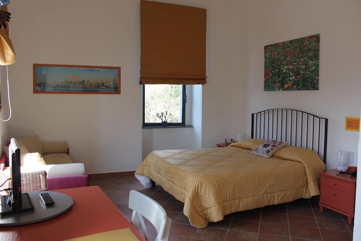 A bedroom with a bed, a desk with a computer, a chair, a window with a view, and a painting on the wall.