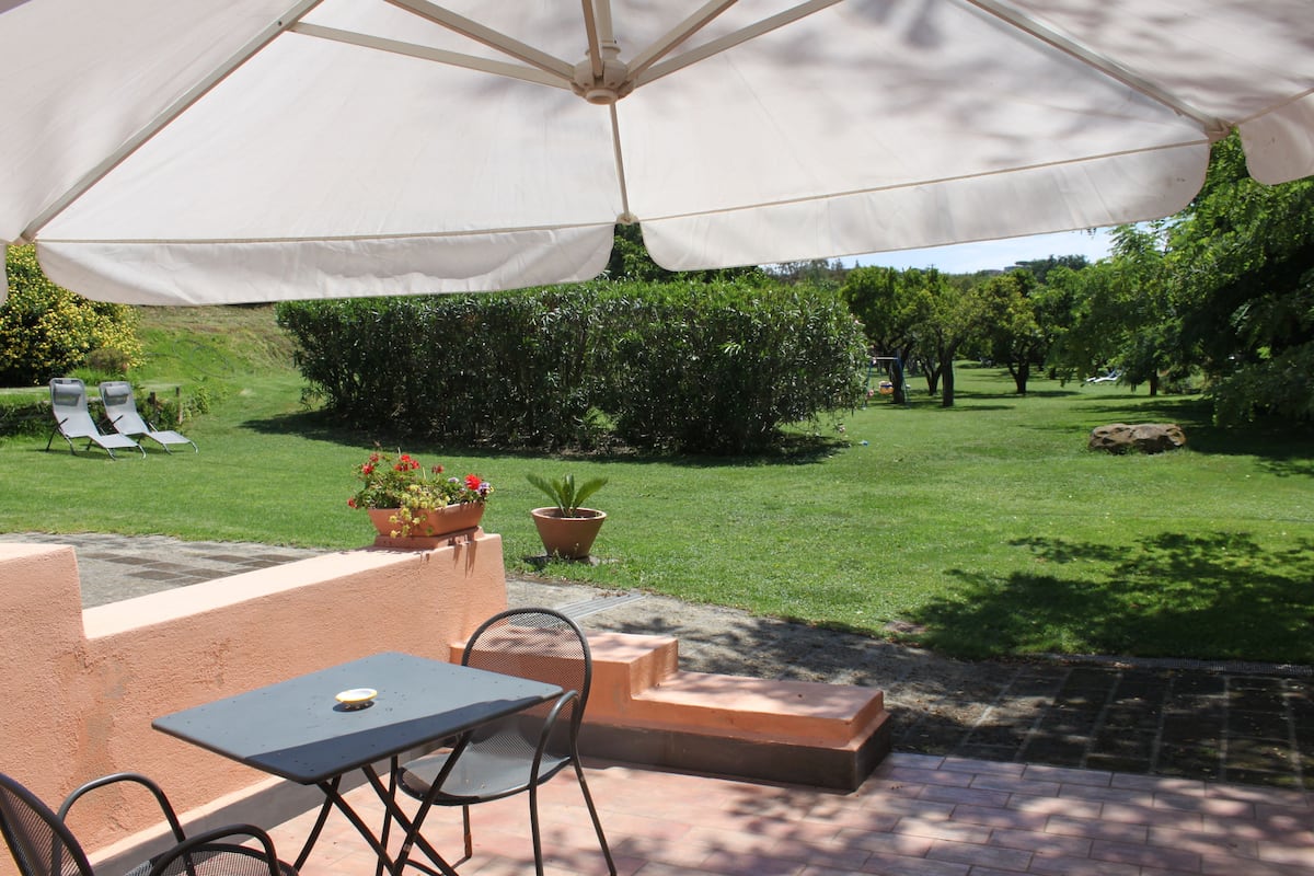 An outdoor patio with a table and chairs under a large umbrella, surrounded by a well-maintained lawn and garden.