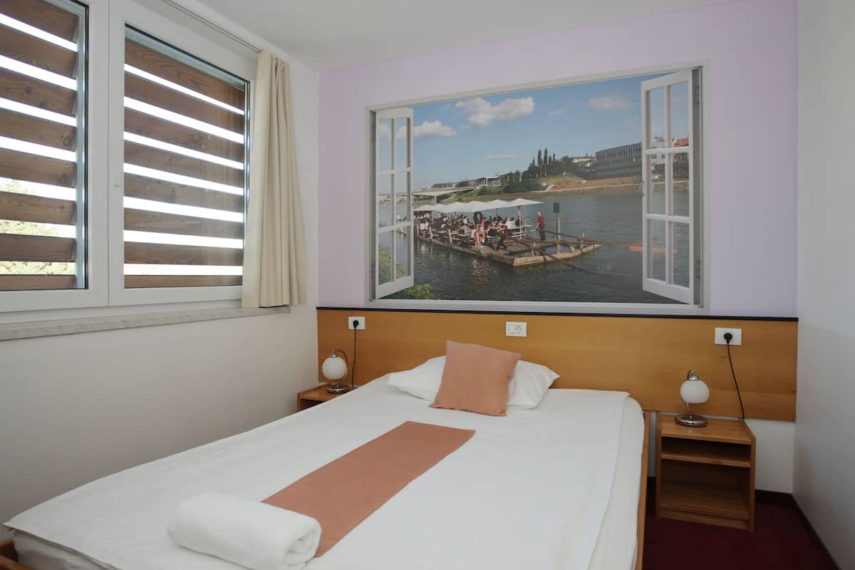 A hotel room with a large bed, a nightstand, a lamp, and a window with blinds.