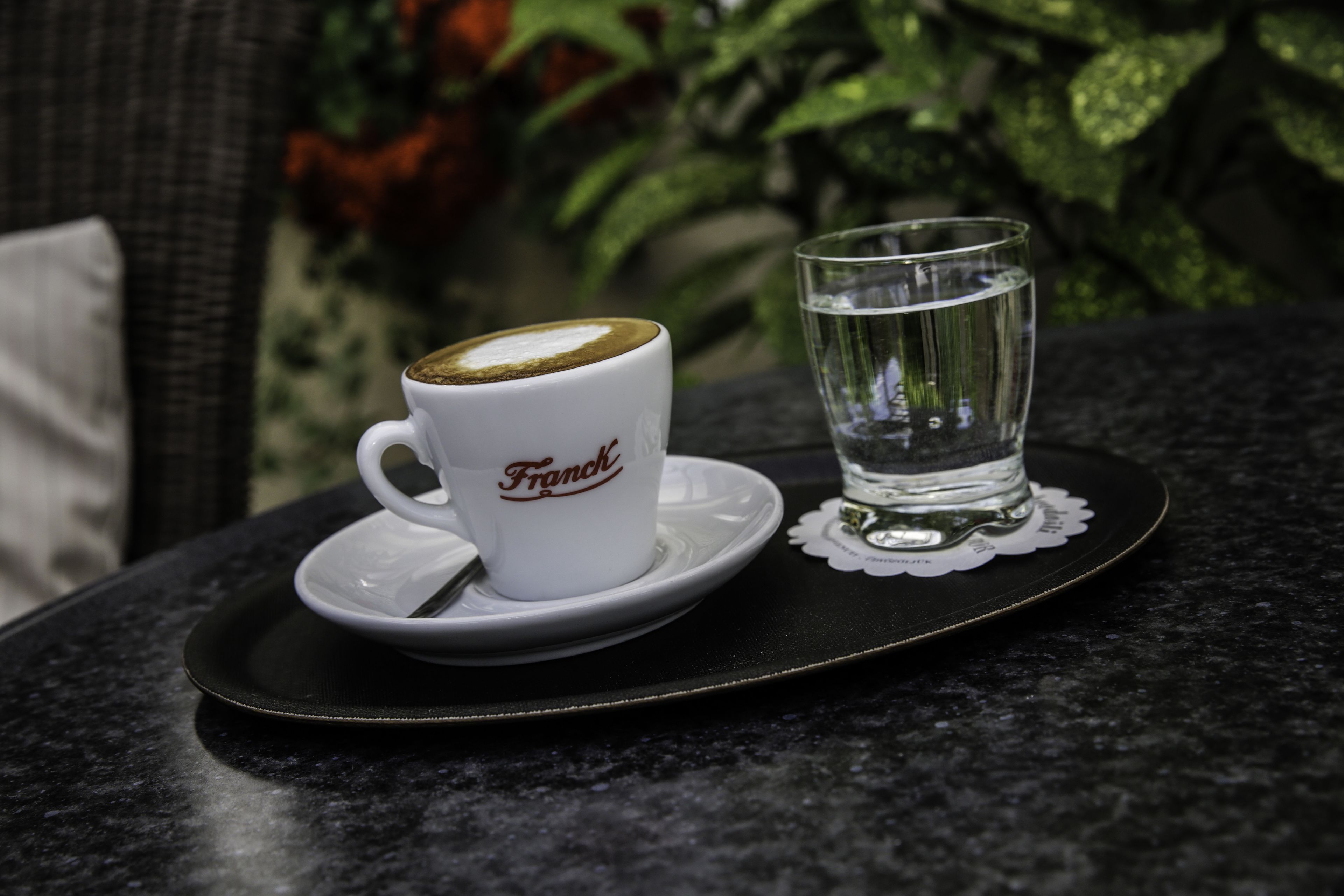 Une tasse à café blanche avec le mot « Franch » écrit dessus, et un verre d’eau transparent sur un plateau noir.